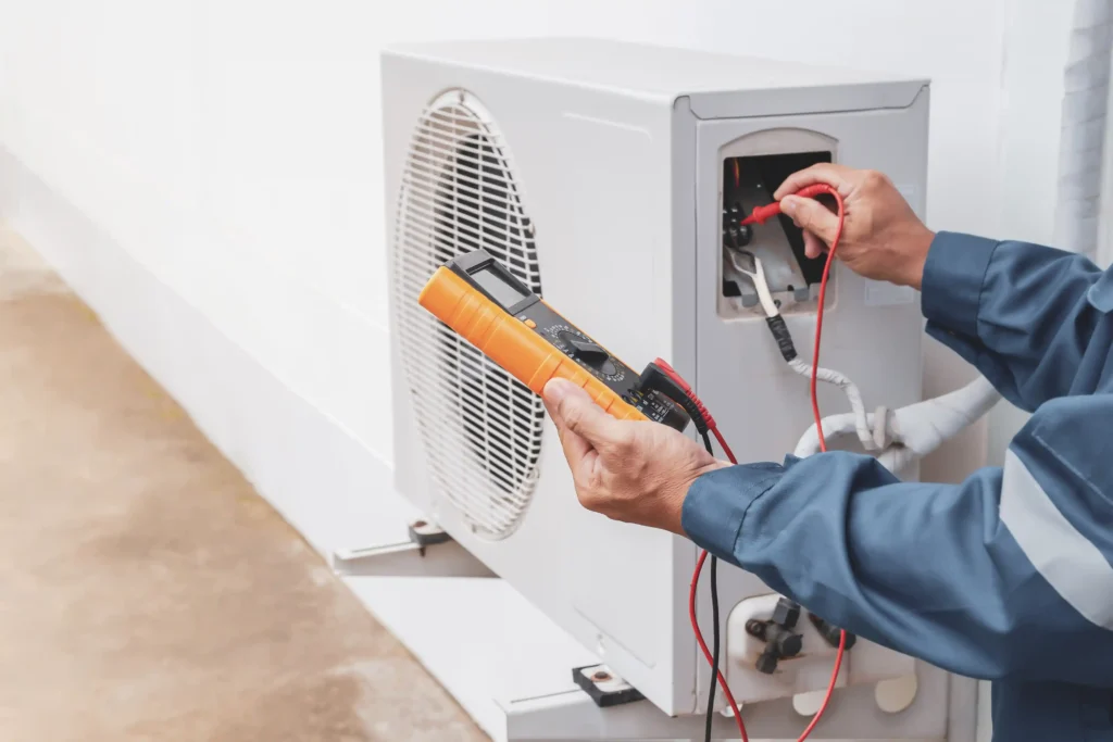 A technician in a blue uniform is using an orange and black digital multimeter to check the electrical connections and voltage inside a white outdoor mini-split AC condenser unit.