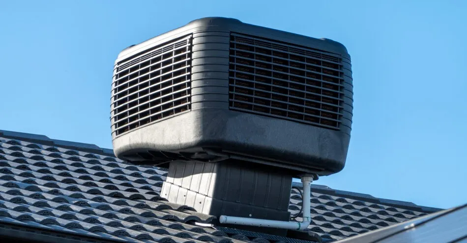 A large, black evaporative cooler unit installed on a dark tiled roof against a clear blue sky.