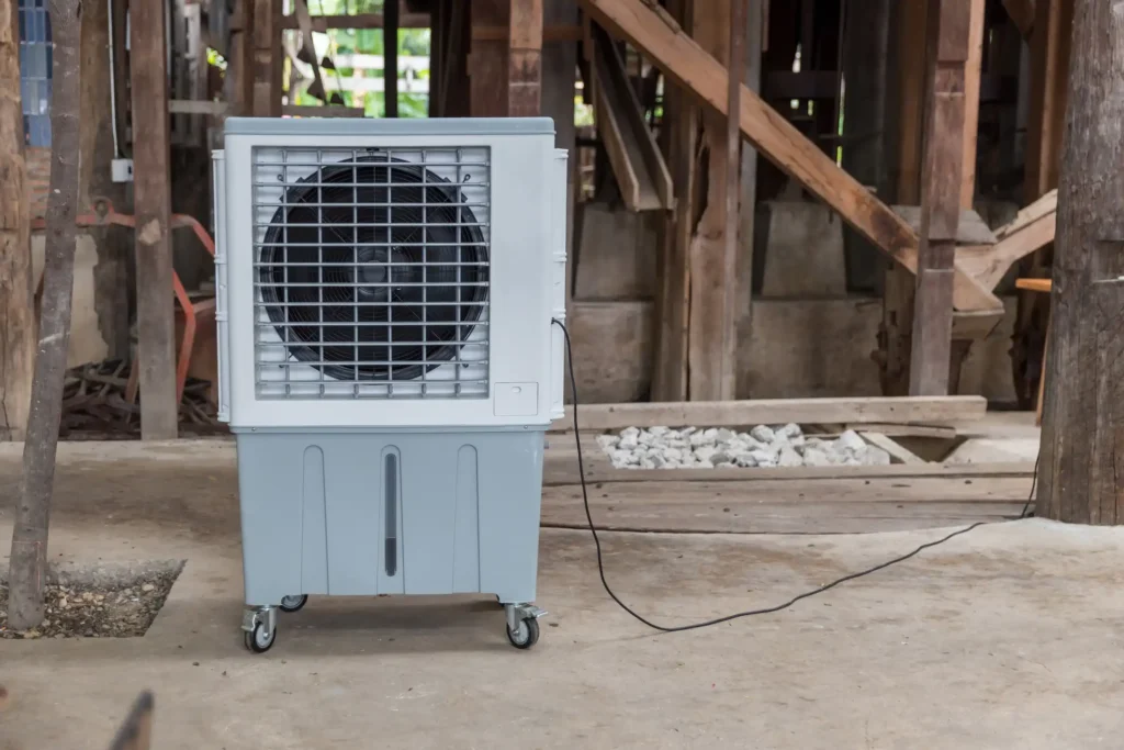 A portable swamp cooler or evaporative cooler with a white top and a grey base, featuring a large black fan grill and caster wheels, sitting on a concrete floor in a rustic, unfinished wooden structure.