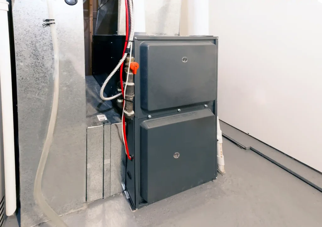 A modern, vertical furnace unit with a dark grey casing and two access panels is installed in a mechanical space with light grey walls and floors. Visible connections include a vertical white plastic exhaust pipe and a black pipe with a red shut-off valve.