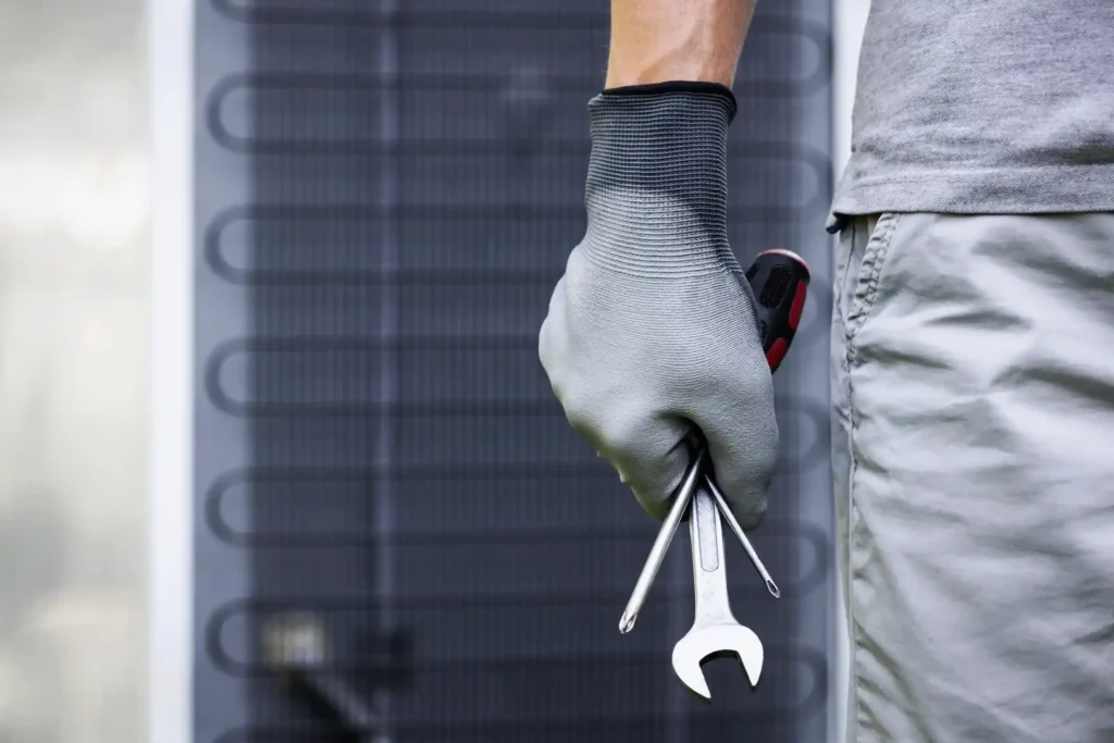 Close-up of a gloved hand holding a wrench and screwdriver in front of an outdoor air conditioning unit.
