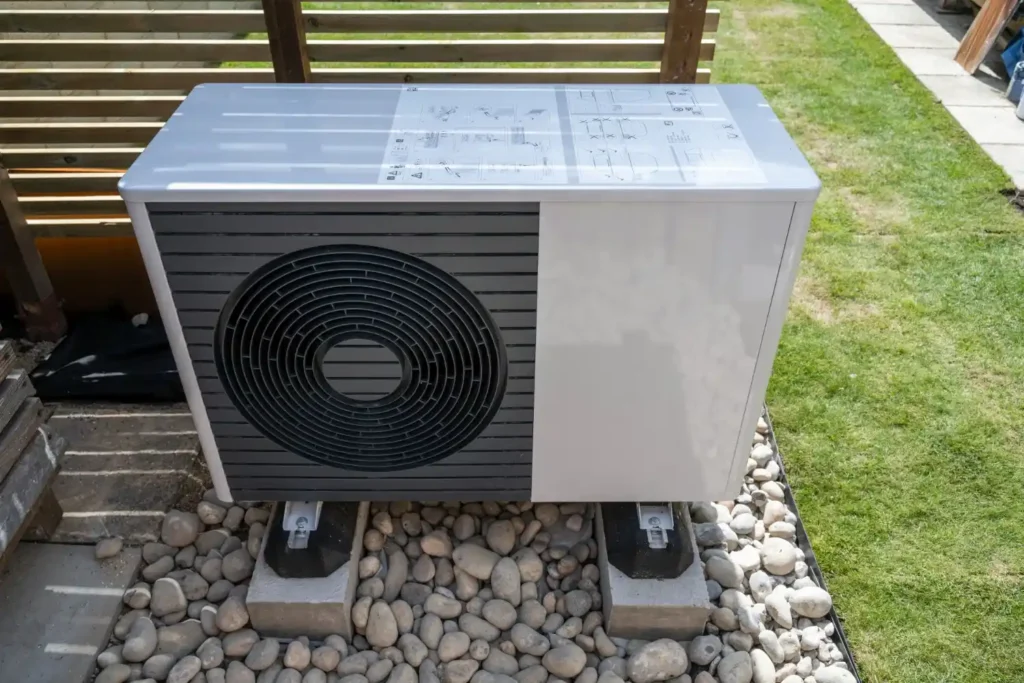 An outdoor heat pump unit resting on concrete blocks on a bed of river stones next to grass.