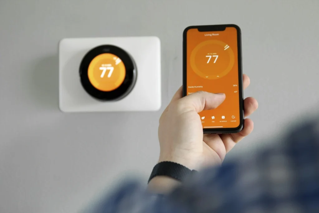 A person holding a smartphone showing a thermostat app next to a wall-mounted smart thermostat.
