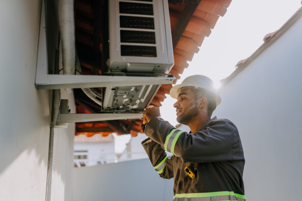 A technician wearing a white hard hat and a dark grey uniform with neon yellow reflective stripes is working on the underside of a white outdoor air conditioning unit that is mounted high on a wall, using a tool, possibly a screwdriver.