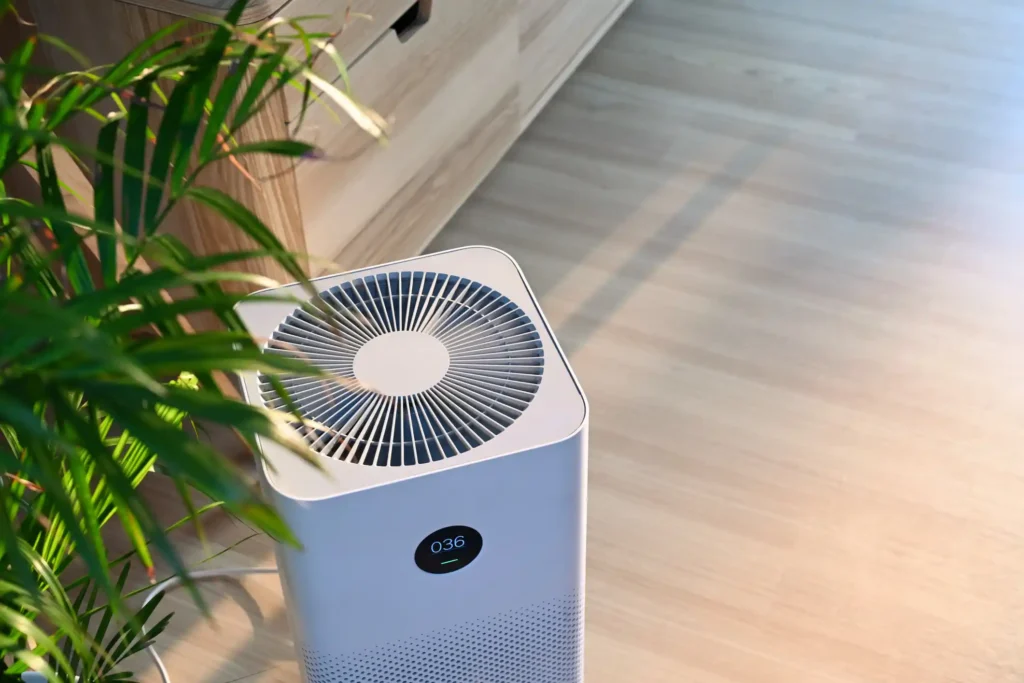 A high-angle, close-up shot of a white, square portable air purifier sitting on a light wood floor next to a green houseplant. The top has a circular grille, and a small digital display on the front shows the number "036".