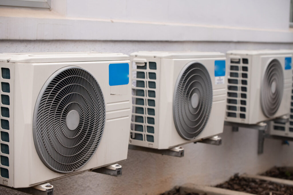 A row of three identical white outdoor air conditioning condenser units is mounted on the wall of a building. Each unit features a large circular front grille with a fan and a small blue sticker on the side.